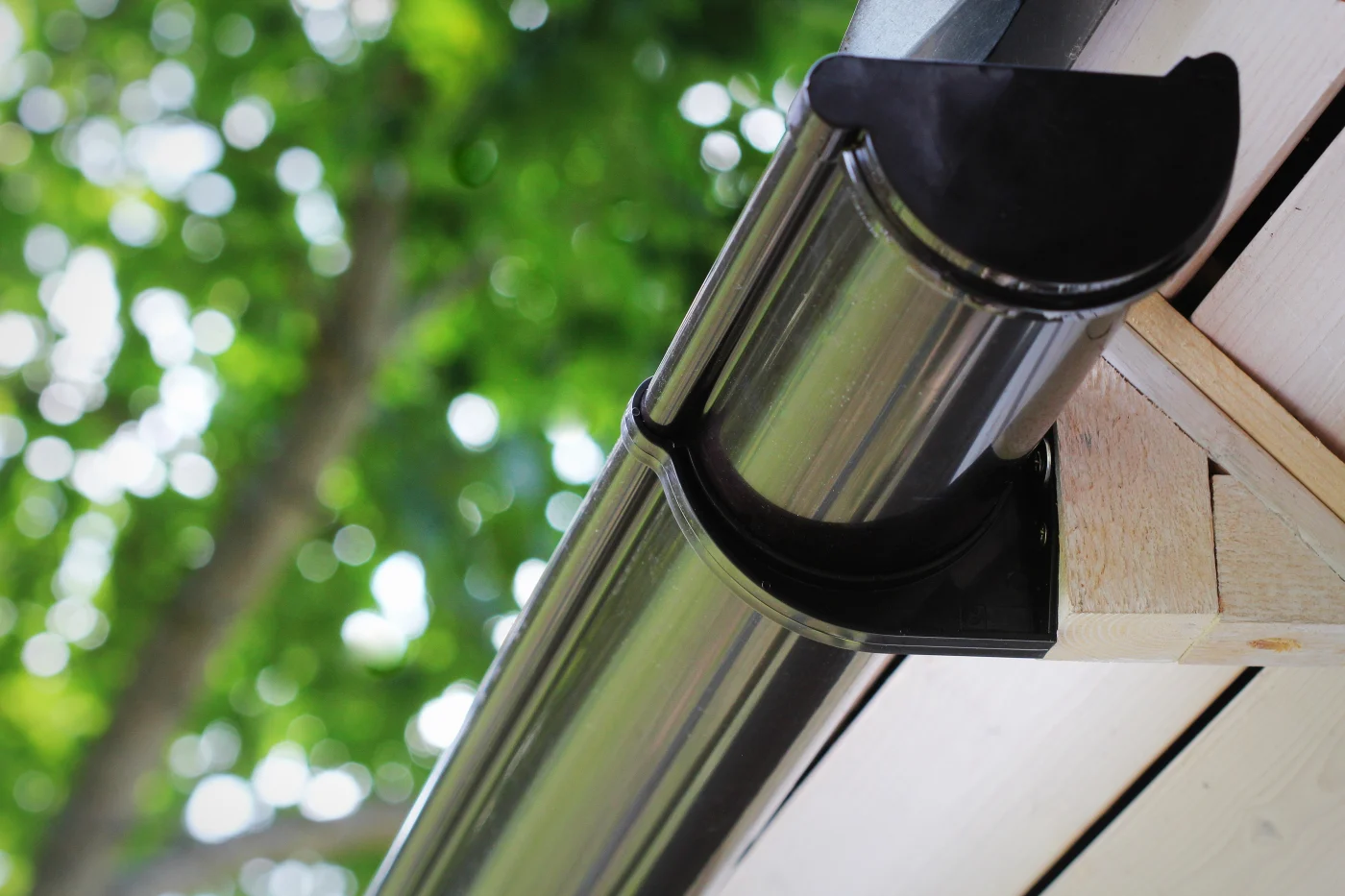 Close-up view of a clean metal rain gutter installed along a light wood roof edge, with lush green trees softly blurred in the background