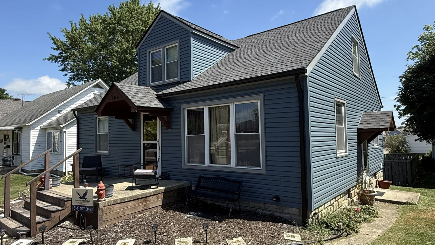 blue two-story home with a gray shingle roof, front dormer, wood porch, and fresh exterior updates on a sunny day.
