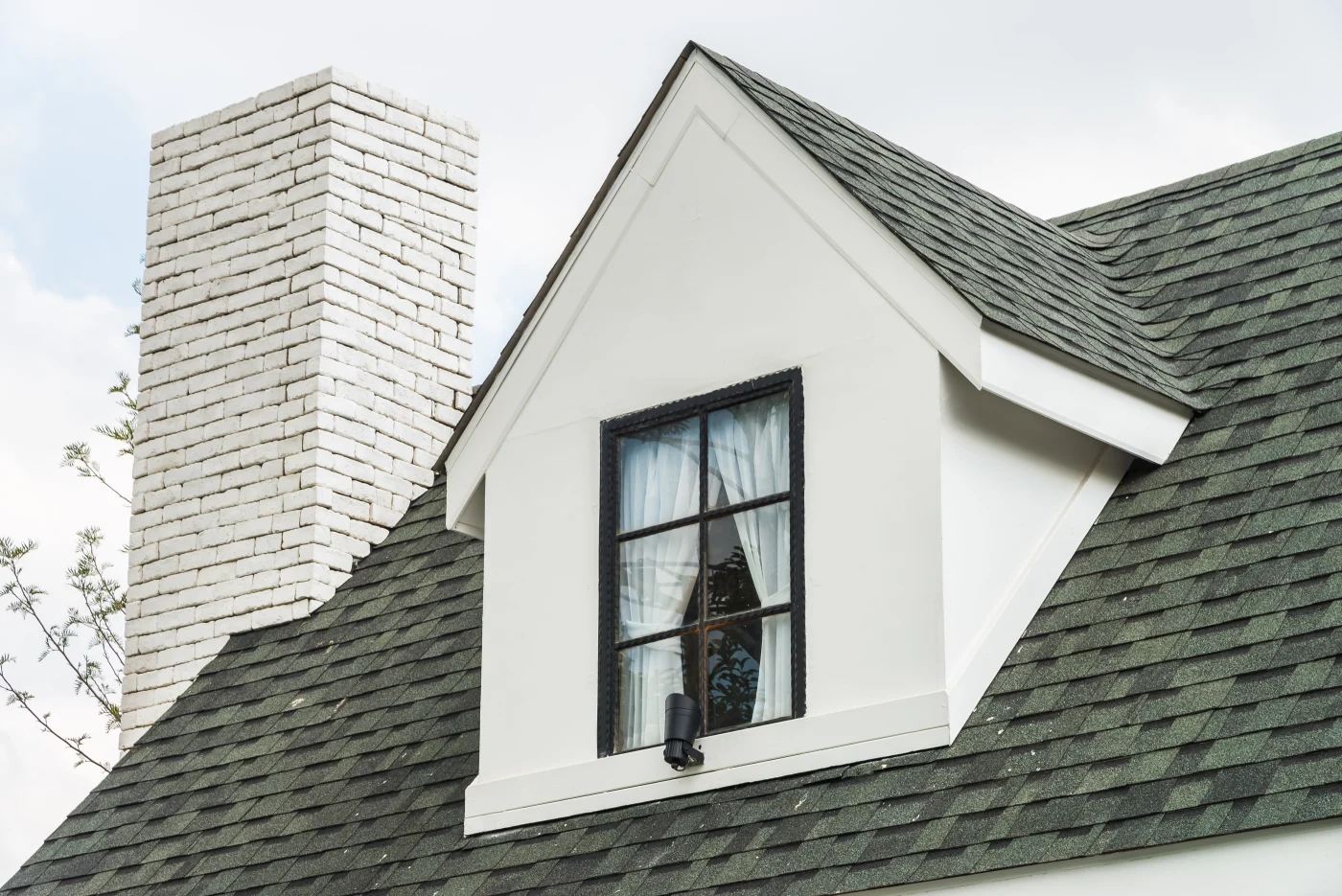 A view of part of a home’s roof, a glass window, and chimney from ground level. The roof is covered in dark colored shingles, the window trim is black, and the chimney is white brick