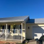 A single-story home with a white garage door and yellow siding. The roof is made of light gray metal, and there is a low front porch that runs along the front of the home. It has a white fence and railing